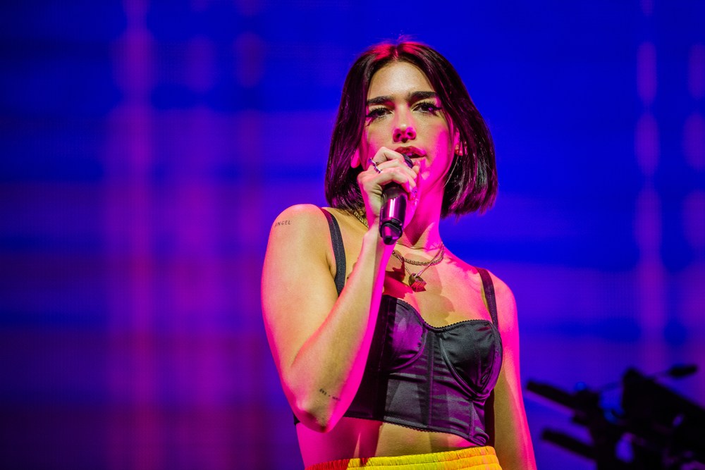 Image of pop star Dua Lipa on stage in a black top with a purple backdrop