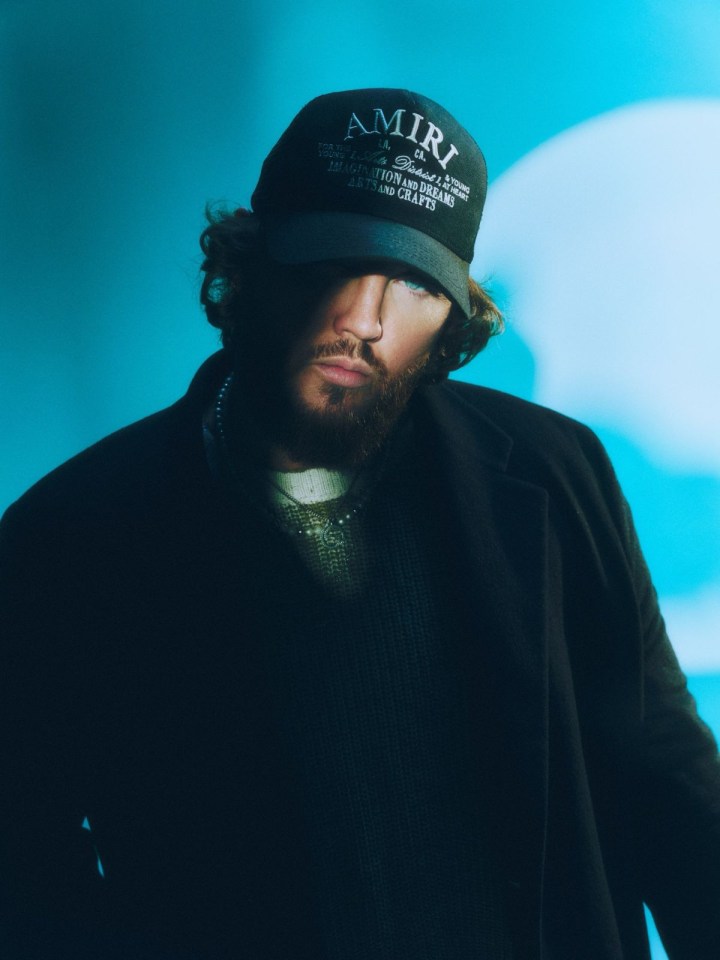 Image of Singer James Arthur in half shadow, half light, wearing dark clothing and a cap that says ‘Amiri’.