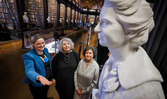 Three women in an old library looking at a white marble sculpture of a woman.